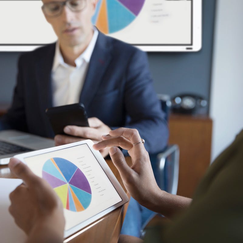 Man in background with whiteboard displaying pie chart and woman holding tablet