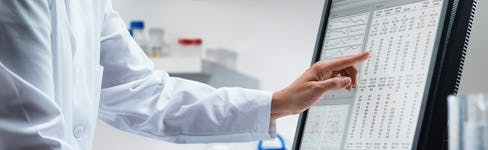 person wearing a lab coat pointing at data on computer screen