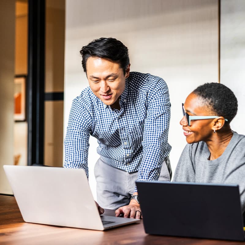 A man and woman, each on their laptop, having a pleasant chat while looking at ways to accelerate speed to market with a service partner.