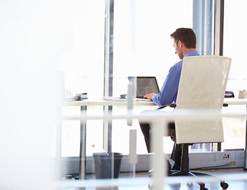 Photo of a man in a blue shirt with his back to the viewer, working on his laptop.