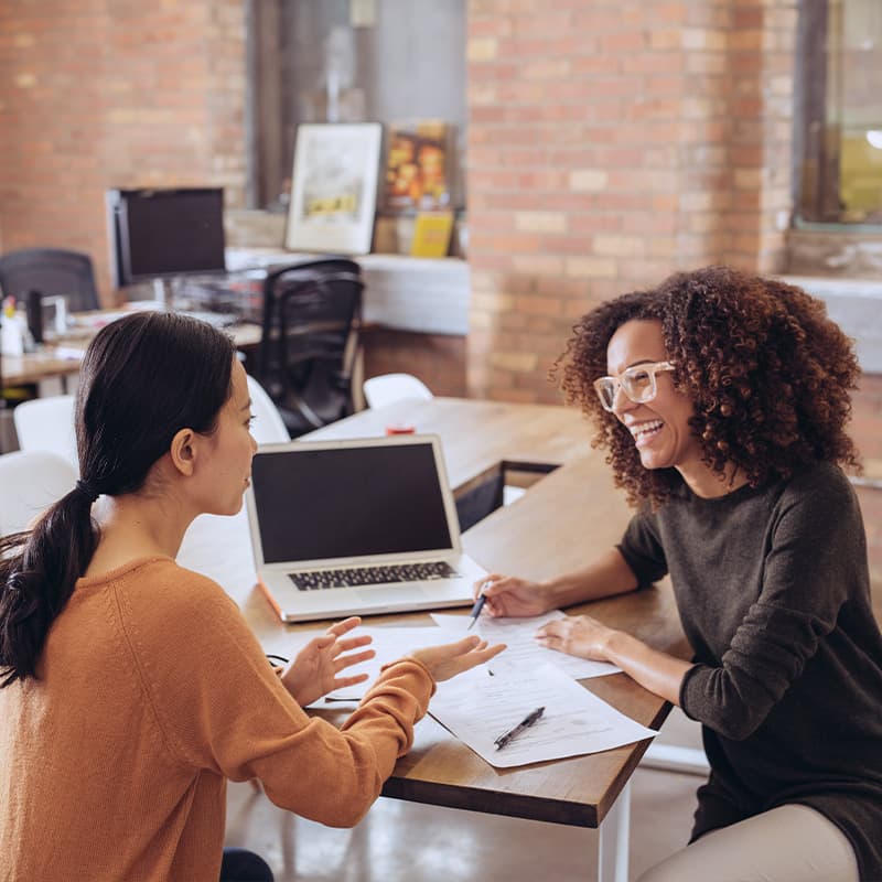 Two women discussing credit union details sitting at a table with papers and laptop on it.