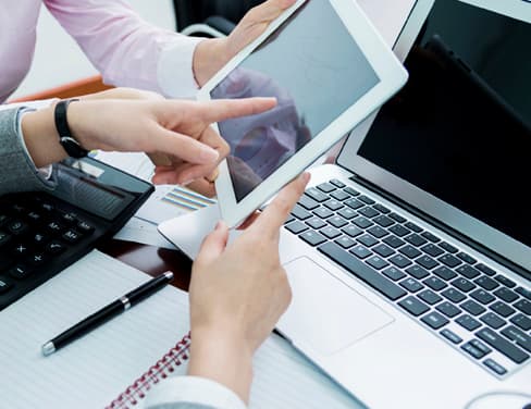 Ricoh hands reviewing tablet over desk with laptop and paper and pen