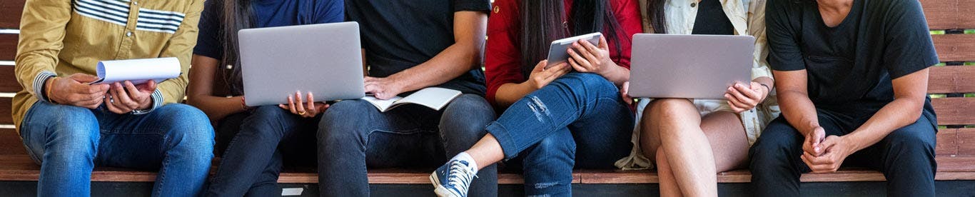 group of college students sitting on bench looking at laptops and tablets, image is cropped to only show mid section of people