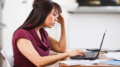 woman sitting at laptop looking frustrated with head in hands and messy desk of papers