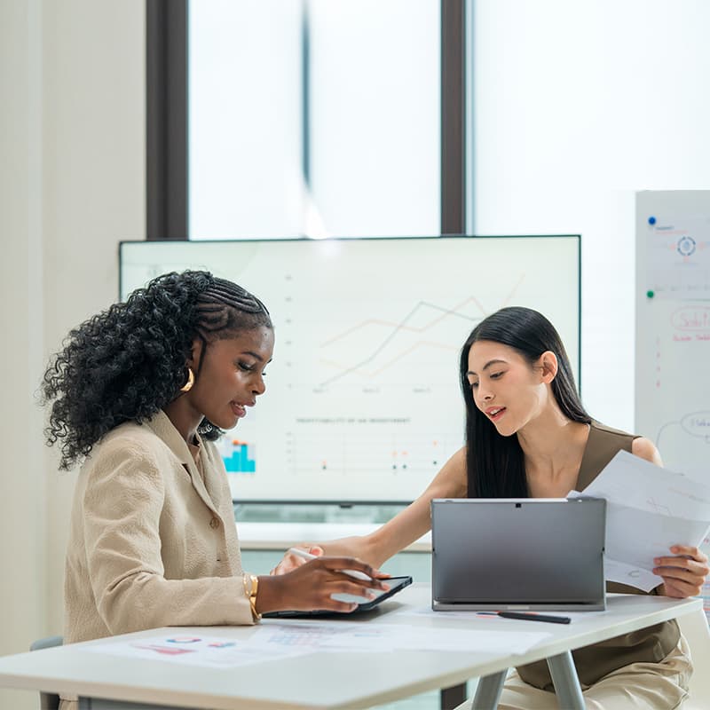Two professional women having a discussion with a laptop in front of them, and a projector with data behind - demonstrating fully managed hospitality services.