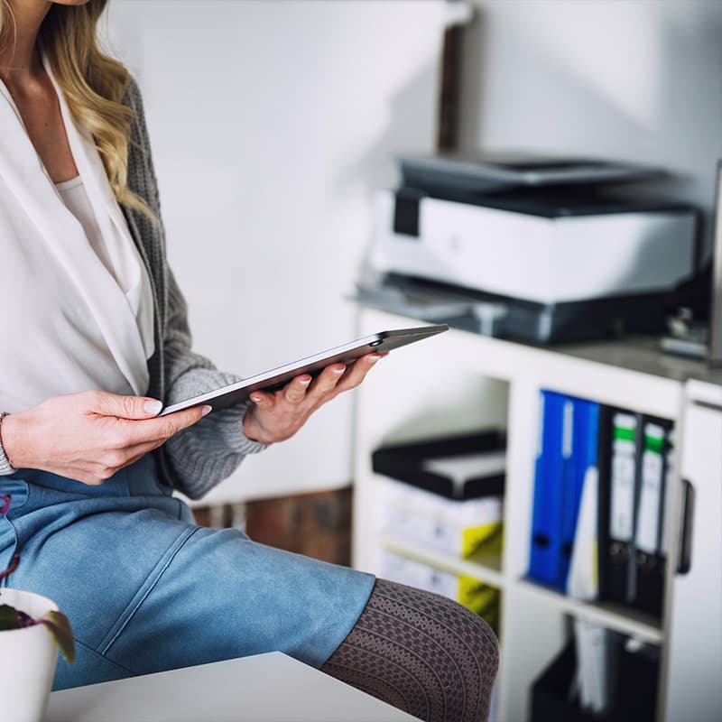 Woman Sitting On Desk Looking At Digital Tablet with printer in the background